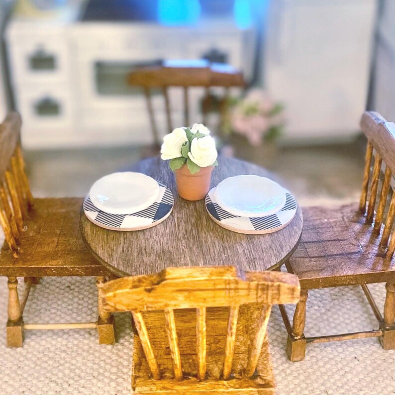 May include: A miniature wooden dining set featuring a round table and three spindle-back chairs. The table is set with two white plates on black and white buffalo plaid placemats and a small terracotta pot containing three white roses.