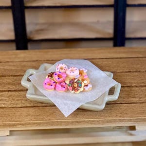 May include: A collection of miniature toy donuts with various colored frostings and sprinkles, arranged on a piece of white parchment paper resting on a small, light-colored rectangular ceramic tray with side handles.