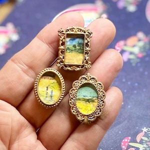 May include: Three miniature landscape paintings in ornate gold frames. The paintings depict fields and skies in yellow, blue, and green hues. The frames are rectangular and oval, with detailed filigree designs. The pieces are held in a hand.