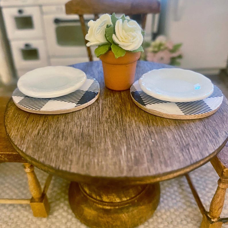 May include: A miniature round wooden table featuring two white plates placed on black and white buffalo plaid coasters. A small terracotta-colored pot sits in the center, containing three white fabric flowers with green leaves.