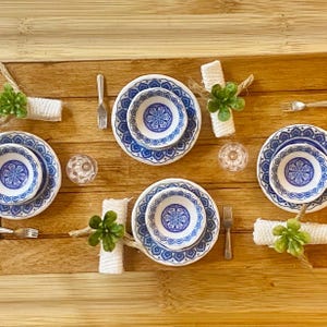 May include: A miniature dining set featuring four white plates and bowls with a blue floral pattern, four silver forks, four clear glasses, and four white napkins wrapped with small green artificial succulents on a wooden surface.