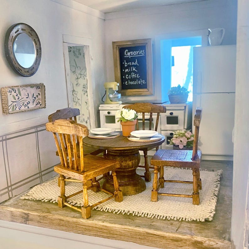 May include: A miniature dollhouse kitchen featuring a round wooden pedestal table with two chairs, a white refrigerator, a yellow stand mixer, and a chalkboard sign with the text Groceries, bread, milk, coffee, and chocolate written on it.