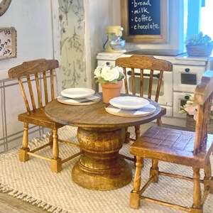 May include: A miniature wooden dining set featuring a round pedestal table and three chairs. The table is set with two white plates on black and white buffalo check placemats and a small terracotta pot containing white flowers.
