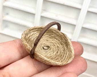 Könnte beinhalten: Ein kleiner gewebter Korb mit einem braunen Griff. Der Korb besteht aus naturbelassenem Garn und hat eine kreisförmige Form. Der Griff besteht aus einem dunkleren braunen Material. Der Korb wird in der Hand einer Person gehalten.