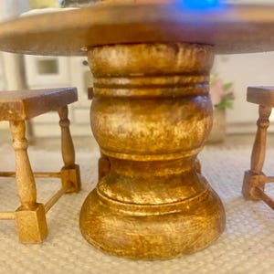 May include: A miniature wooden dining set featuring a round table with a thick, pedestal-style base and two matching benches with slender, turned legs. The furniture is crafted from light brown wood with a visible grain and smooth finish.