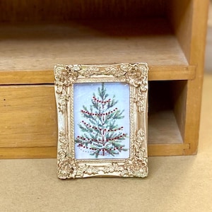 May include: A small, ornate gold-coloured picture frame with a miniature painting of a Christmas tree decorated with red baubles. The frame has a detailed, textured design. The painting is set against a light blue background.