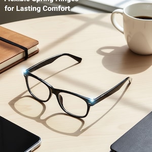 May include: Black-framed eyeglasses with clear lenses rest on a light-colored wooden surface. The glasses are next to a smartphone, a coffee mug, and a closed notebook. The text "Flexible Spring Hinges for Lasting Comfort" is at the top.