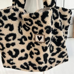 Könnte beinhalten: Eine beige und schwarze Tasche mit Leopardenmuster und einem kleinen schwarzen Herz-Patch.