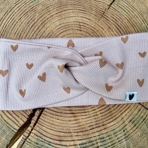 Könnte beinhalten: Ein helllila Stirnband mit gerippter Struktur und einem Twist-Design. Das Stirnband ist mit kleinen braunen Herzen verziert. Ein kleines Etikett mit einem schwarzen Herzen ist angebracht. Das Stirnband ist aus weichem Stoff.