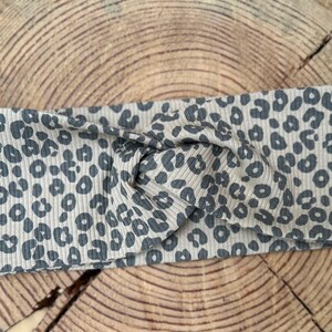 Könnte beinhalten: Ein graues und weißes Stirnband mit Leopardenmuster und einem verdrehten Knotendetail. Das Stirnband besteht aus einem gerippten Stoff und liegt auf einer Holzoberfläche. Das Tiermuster ist ein sich wiederholendes Design aus dunkelgrauen Flecken.