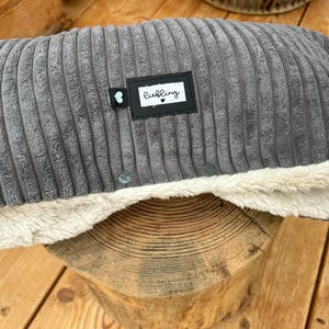 Könnte beinhalten: Ein graues Cord-Haustierbett mit einem cremefarbenen, plüschigen Innenraum. Das Bett hat ein schwarzes Etikett mit dem Wort "liebling" in Weiß. Das Bett liegt auf einer Holzoberfläche.