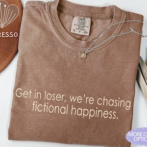 May include: Espresso-colored t-shirt with the text "Get in loser, we're chasing fictional happiness." A silver necklace with a pendant rests on the shirt. The shirt has a crew neck and short sleeves.