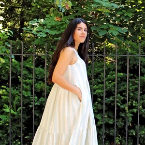 Può includere: Abito lungo senza maniche a righe verticali in azzurro e bianco. L'abito ha una gonna a balze e tasche. La persona indossa sandali dorati. Lo sfondo comprende una recinzione in metallo nero e fogliame verde.