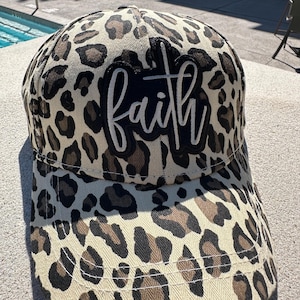 May include: A baseball cap with a leopard print pattern in shades of tan, brown, and black. The cap features the word "faith" in white script on a black patch. The cap has a curved brim.