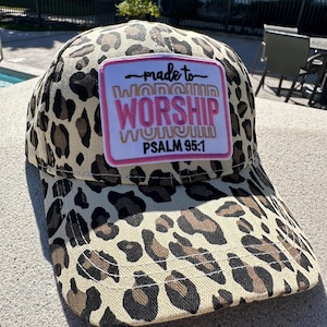 May include: A baseball cap with a leopard print pattern in tan, brown, and black. The cap features a rectangular patch with the words "made to WORSHIP" and "PSALM 95:1" in pink and gold lettering.