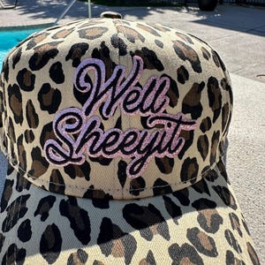 May include: A baseball cap with a tan and brown leopard print pattern. The cap features the text "Well Sheeeyit" in a pink glittery script across the front. The cap is made of a durable fabric.