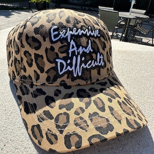 May include: A baseball cap with a tan and brown leopard print pattern. The cap features the embroidered text "Expensive And Difficult" in white thread. The hat has a curved brim and a classic six-panel design.