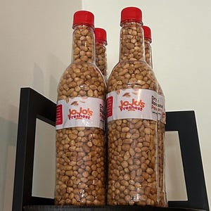 May include: Four clear plastic bottles filled with peanuts, each topped with a red cap. The bottles have labels that read "JoJo's Freshest" and "Dry Roasted Peanuts". The bottles are arranged on a black shelf.