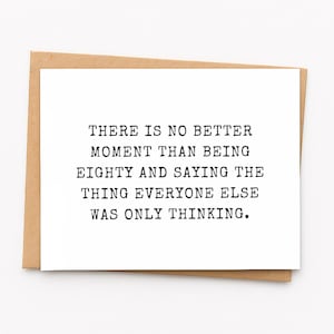 May include: A white rectangular greeting card with black typewriter-style text that reads: THERE IS NO BETTER MOMENT THAN BEING EIGHTY AND SAYING THE THING EVERYONE ELSE WAS ONLY THINKING. The card is placed on top of a brown kraft paper envelope.