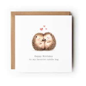 May include: A square white greeting card with a brown envelope behind it. The card features an illustration of two brown hedgehogs hugging with three small pink hearts above them. The text reads: Happy Birthday to my favorite cuddle bug.
