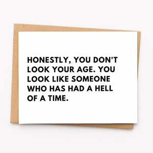May include: A white greeting card with black text that reads: HONESTLY, YOU DON'T LOOK YOUR AGE. YOU LOOK LIKE SOMEONE WHO HAS HAD A HELL OF A TIME. The card is placed on top of a light brown kraft paper envelope, showing a simple rectangular design.
