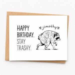 May include: A white greeting card with a brown envelope featuring a black ink illustration of a raccoon. The text reads: HAPPY BIRTHDAY. STAY TRASHY. Above the raccoon, the name jimothy is written in lowercase letters between two small hearts.