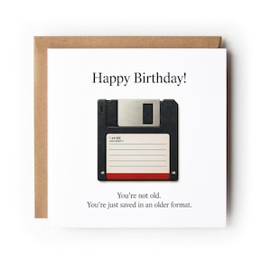 May include: A square white greeting card featuring a black floppy disk illustration in the center. The text reads Happy Birthday! You're not old. You're just saved in an older format. The card is paired with a brown envelope behind it.