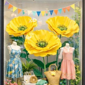 May include: A storefront display featuring three large yellow poppy flowers. Two dresses on mannequins are visible, one blue floral and one pink. A straw bag, white shopping bags, and other accessories complete the summer-themed display.