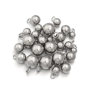 Peut inclure: Une collection de breloques sphériques en métal argenté, chacune dotée d'un petit anneau de jonction circulaire fixé sur le dessus. Les sphères ont une surface lisse, réfléchissante et polie, et varient en taille, formant un amas de perles.