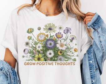 May include: White oversized t-shirt with a floral print and the text "GROW POSITIVE THOUGHTS" in brown lettering.