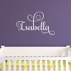 May include: White vinyl wall decal with the name "Isabella" in a cursive font with a decorative swirl design. The decal is displayed above a white crib with a yellow and white striped mattress.