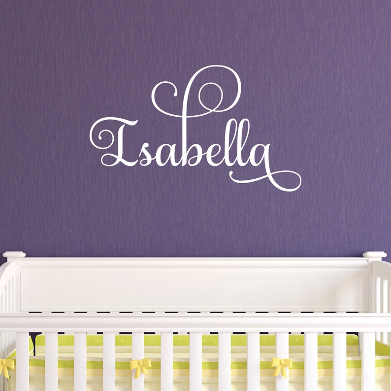 May include: White vinyl wall decal with the name "Isabella" in a cursive font with a decorative swirl design. The decal is displayed above a white crib with a yellow and white striped mattress.
