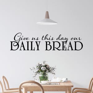 May include: A wall decal with the text "Give us this day our DAILY BREAD" in black script. The decal is displayed above a wooden table with a vase of flowers and two chairs. A modern light fixture hangs from the ceiling.