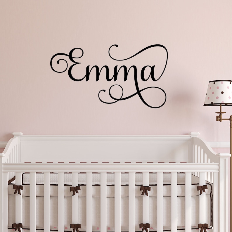 May include: A white crib with a brown and white patterned bumper pad. The wall behind the crib is a light pink color and has a black vinyl decal with the name "Emma" in a cursive font.