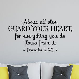 May include: Black text on a gray wall reads "Above all else, GUARD YOUR HEART, for everything you do flows from it. ~ Proverbs 4:23~". The text is above white pillows with decorative pillows.