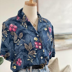May include: A dark blue floral shirt featuring pink and yellow flowers and beige leaves. The shirt is worn with light blue denim jeans and a black belt. The outfit is displayed on a mannequin.