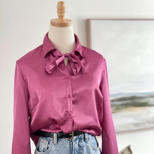 May include: A magenta satin blouse with a bow tie collar. The long-sleeved shirt is buttoned down the front and is paired with blue jeans and a black belt. The blouse is displayed on a mannequin.