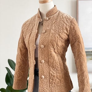 May include: A tan quilted jacket with a floral pattern and a mandarin collar. The jacket has a button-up front and long sleeves. The jacket is displayed on a mannequin.