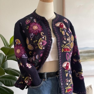 May include: A dark navy blue long-sleeve cardigan featuring intricate, multi-coloured floral embroidery in shades of magenta, yellow, and green. The garment has a round neckline, a button-front closure, and visible whipstitch detailing along the edges.