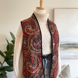 May include: A sleeveless waistcoat featuring a vibrant red, black, and gold paisley pattern with black trim. The waistcoat is worn over a cream-coloured cable knit turtleneck jumper and light blue denim jeans, secured with a black leather belt.