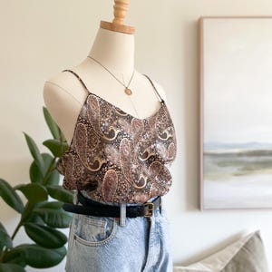 Peut inclure: Un haut caraco sans manches avec un imprimé cachemire marron, noir et crème, doté de fines bretelles, associé à un jean en denim bleu clair et une ceinture en cuir noir avec une boucle dorée, présenté sur un mannequin.