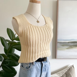 May include: A sleeveless, cream-coloured ribbed knit top with a square neckline, worn with light blue denim jeans and a black leather belt. A thin gold-toned necklace with a small circular pendant rests against the neckline of the knit garment.