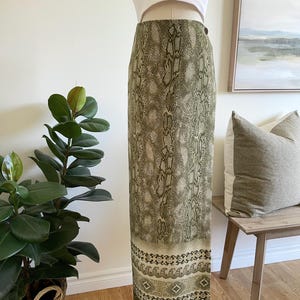 May include: An olive green skirt featuring a snakeskin print. The skirt has a decorative border with geometric and floral patterns in beige and olive green. The skirt is displayed on a mannequin.