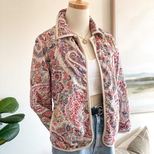 May include: A quilted jacket with a cream base and a vibrant paisley pattern in red, blue, and green. The jacket has a collared neckline, button closure, and cream trim. The jacket is displayed on a mannequin.