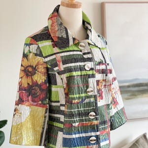 May include: A patterned jacket with a floral and geometric design. The jacket has a collared neckline, three-quarter sleeves, and large, round buttons. The colour palette includes yellow, green, red, and white.