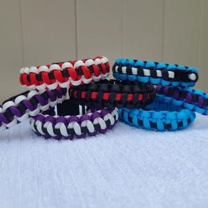 May include: A collection of braided paracord bracelets in various colors, including red, white, black, blue, and purple. The bracelets are arranged on a white surface, showcasing their intricate woven patterns. These accessories are suitable for everyday wear.