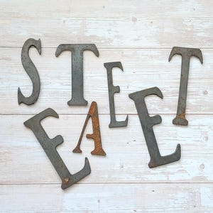 May include: Metal letters spelling out the word "STREET" in a rustic style. The letters are painted in a dark gray color and have a slightly distressed finish. The letters are arranged on a white wooden background.