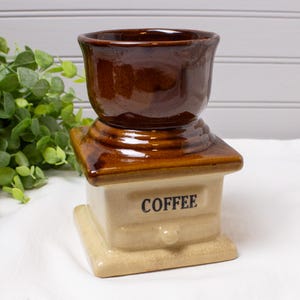 May include: A vintage ceramic coffee grinder with a brown top and a tan base. The word "COFFEE" is printed in black on the front. The grinder has a classic, rustic design, perfect for kitchen decor.