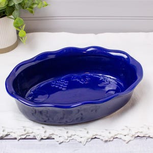May include: A deep blue ceramic oval baking dish with a scalloped rim. The dish features embossed grape cluster designs on the interior. The dish is sitting on a white surface.