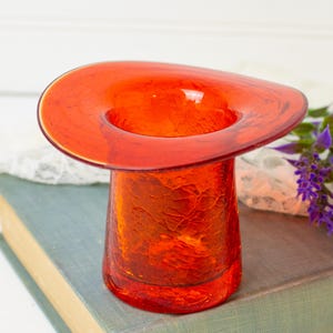 May include: A vibrant orange glass vase featuring a distinct crackle texture throughout its cylindrical body and a wide, flared, circular rim. The glass is translucent, allowing light to pass through the deep orange hue of the material.